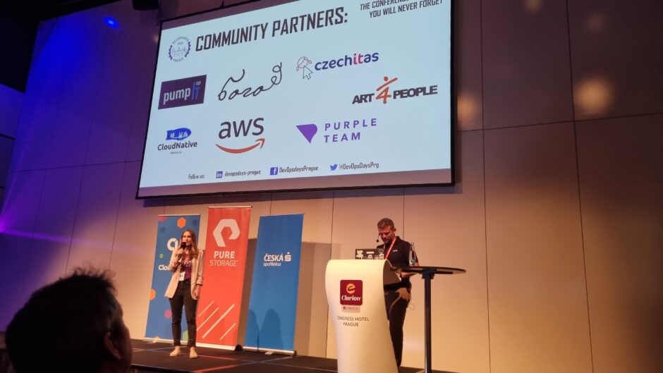 A man and a woman below a huge screen with the community sponsors of the DevOpsDays 2023 Prague