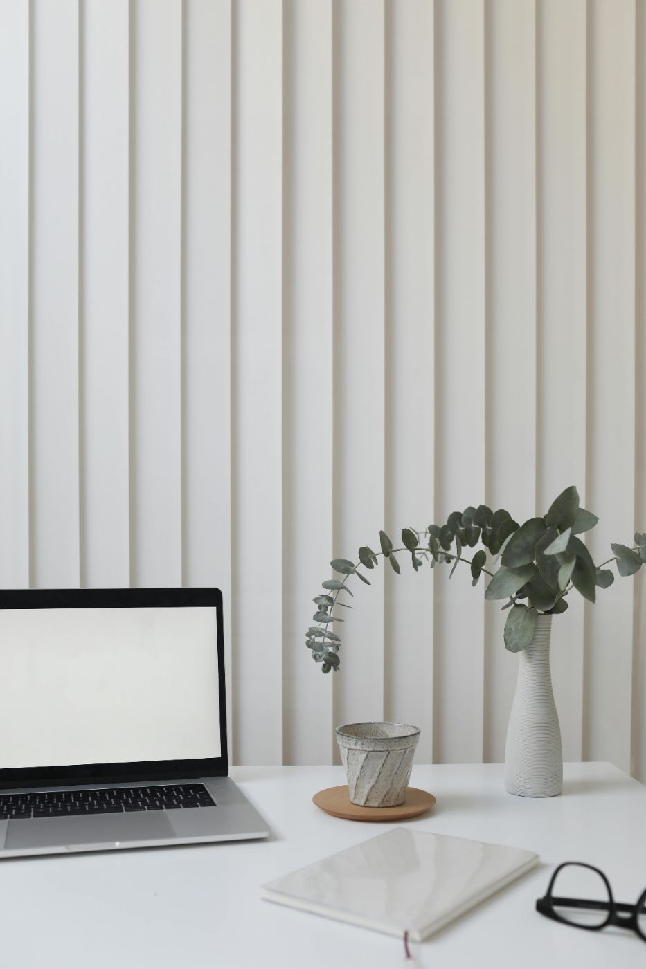 A desk with a laptop, cup, flower, notebook, and glasses.