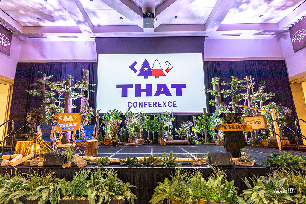 The stage at THAT conf with the logo on the screen, and the whole stage is covered with fake plants, trees, and stumps.