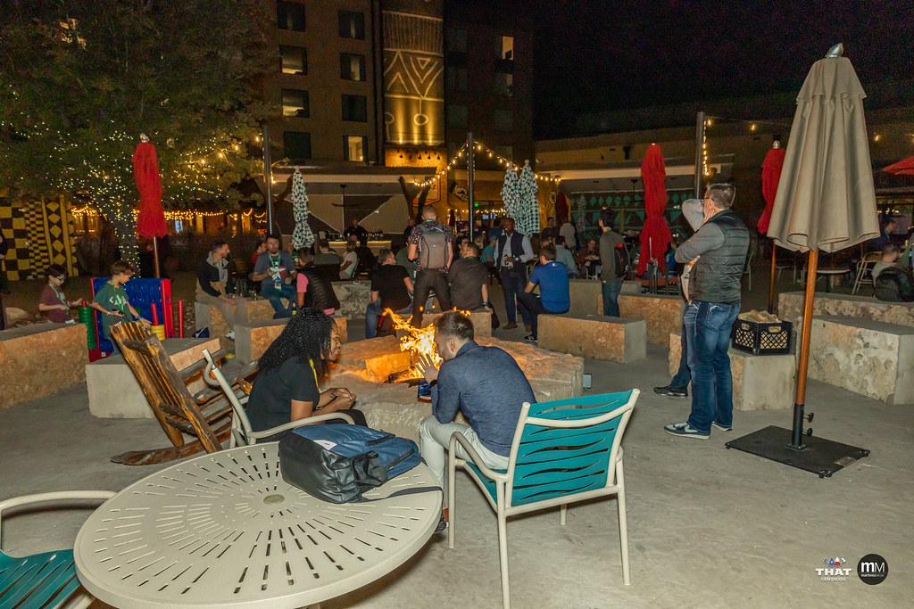 Several people sitting outside at night in a courtyard. There is a fire pit and some string lights. It's very cozy.