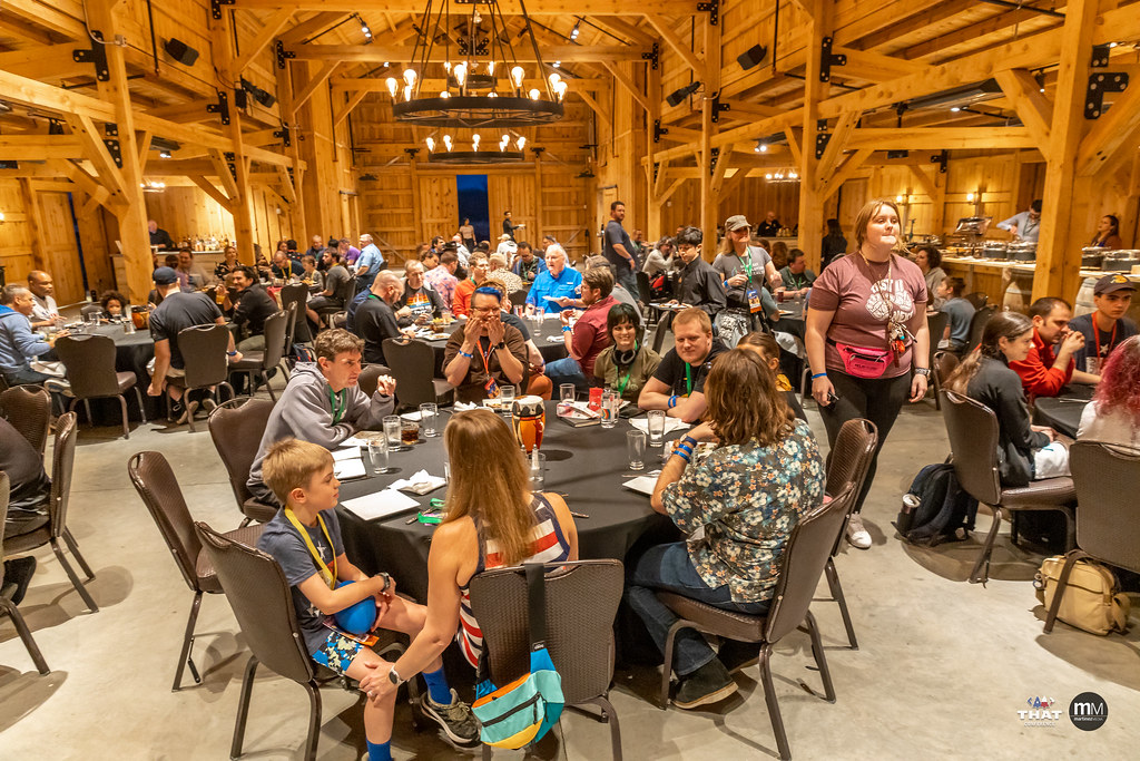 Lots of people sitting around tables in a well-lit barn eating a meal and chatting.
