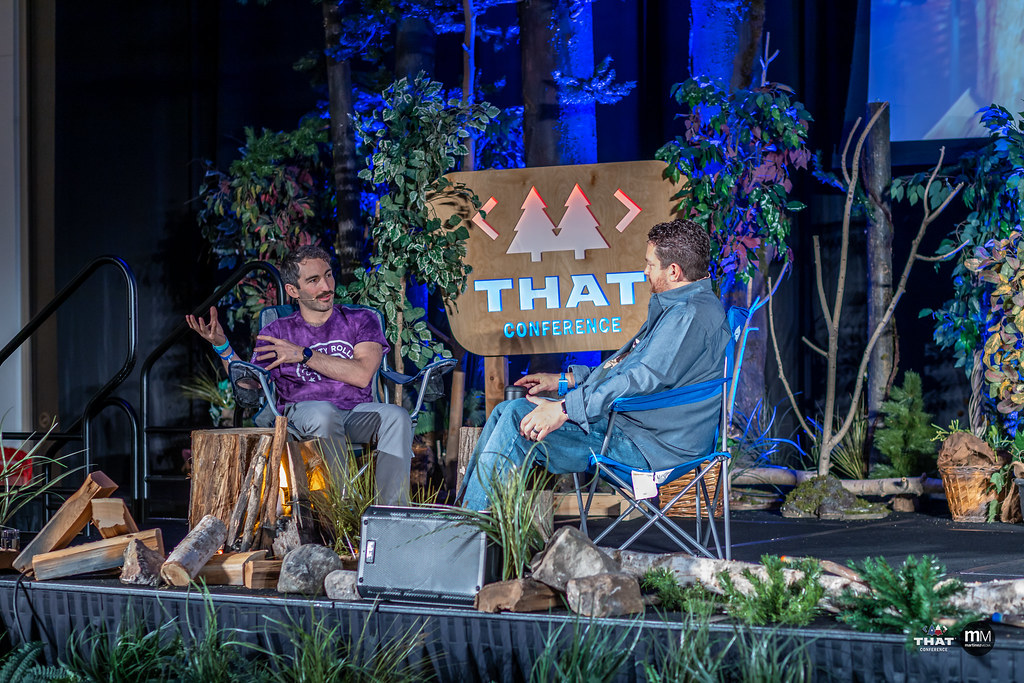 Me sitting with Clark in some camping chairs on the conference stage. We're recording an episode of the podcast, and we're surrounded by fake trees, plants, logs, and rocks.